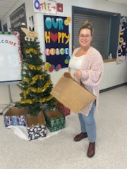 alt="Sarah standing in front of Christmas tree in Bowden Grandview school with a paper bag of donations"